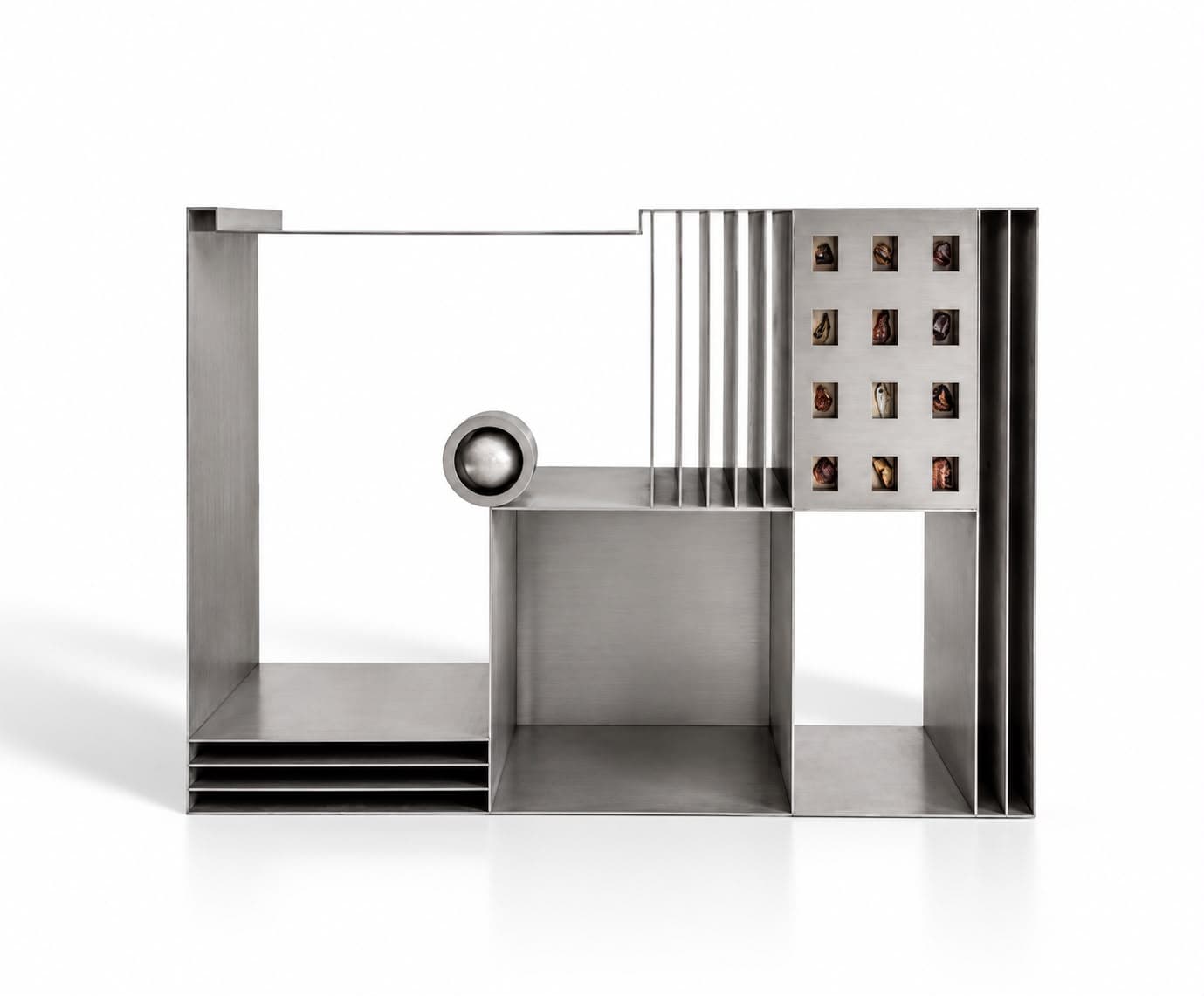 A wide low steel unit of open bays: stacked flat shelves at the left, a polished steel sphere set into a socket at the centre, upright fins for records, and a grid of twelve small specimens framed into the right-hand panel