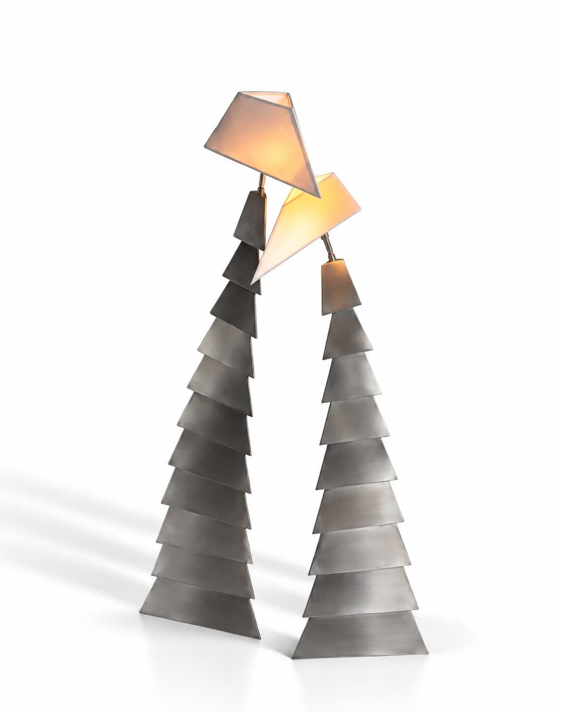 Two stacked lamp towers of tapering blackened steel plates, one taller than the other, each leaning slightly inward and carrying an angular lit shade on a thin stem