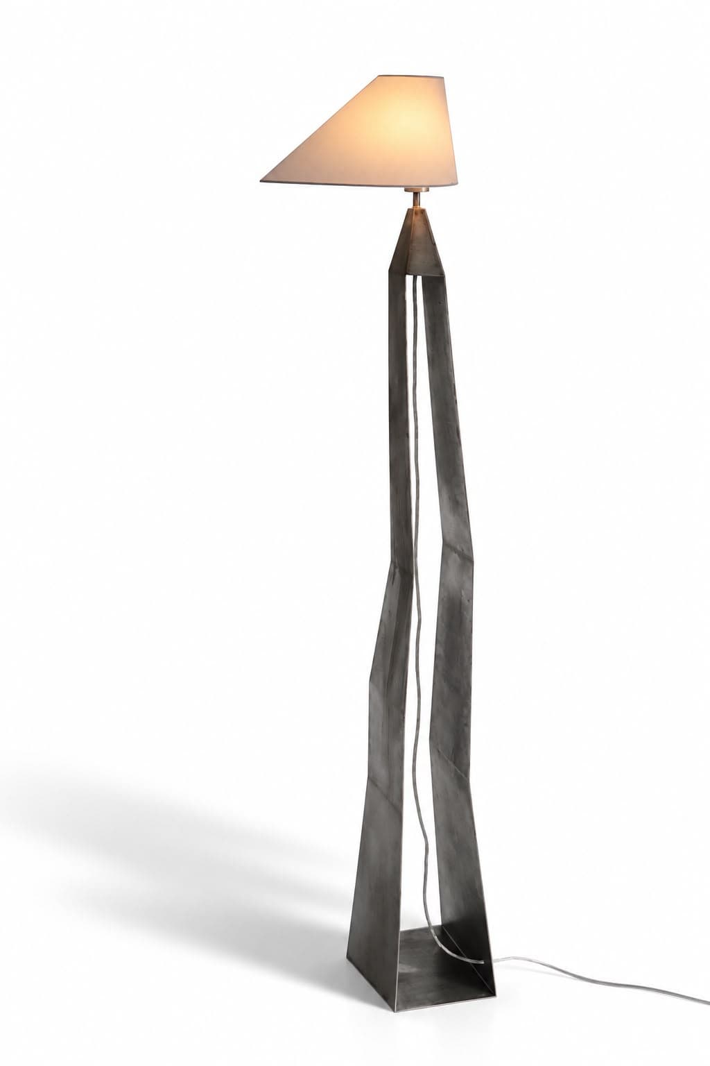 A tall slender floor lamp: two tapering blades of blackened steel rise from a small triangular base plate, kink inward halfway up and meet at a point near the top, where a thin stem carries a wide cream conical shade lit from within and tilted forward; the cable runs down between the blades and trails away across the floor