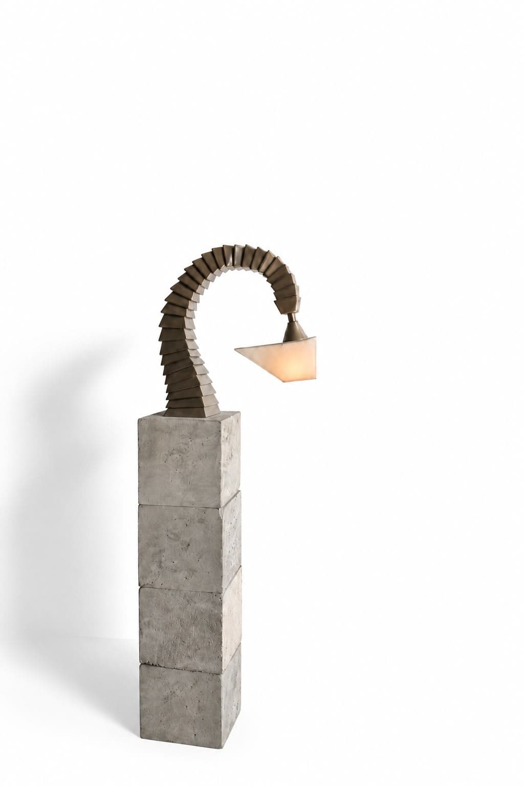 A lamp standing on a column of four stacked concrete blocks: a neck of overlapping tapering steel plates curves up from the top of the column and arches over, ending in a short stem that carries a small downturned triangular shade lit warm from inside