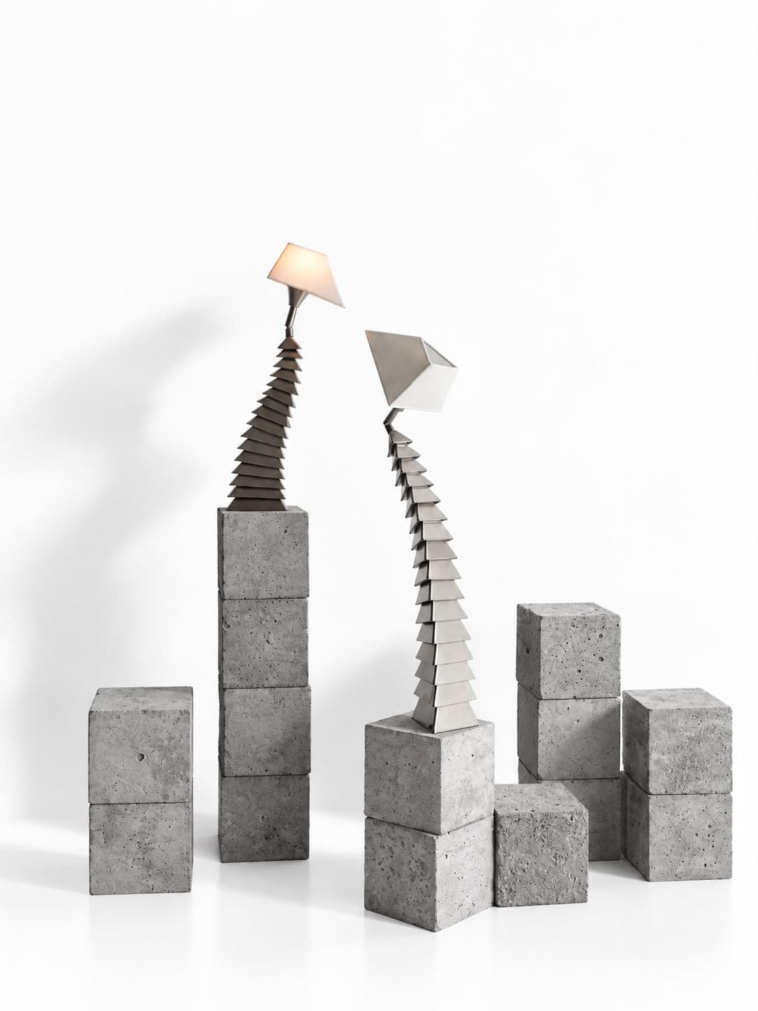 Two of the same lamps closer to, one on a tall column of four concrete blocks under a warm-lit shade, the other on a lower column with its steel shade tipped open toward it, with single and paired concrete blocks standing loose across the floor around them