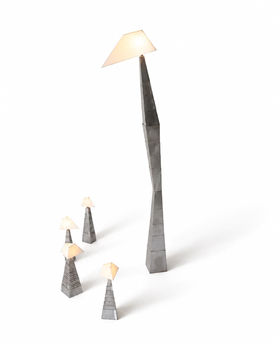 Five lamps of one family standing apart on a white floor: a tall leaning column of stacked tapering steel segments carries a wide pale shade lit from within, and four much smaller lamps stand grouped at its foot, each a stepped steel pyramid under its own small shade
