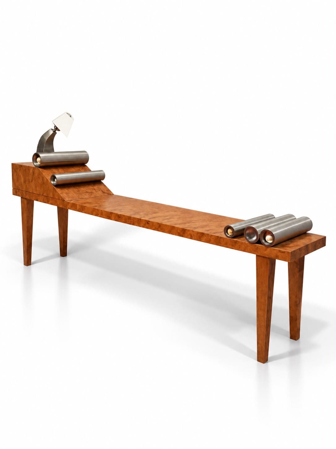 A long low bench in figured rosewood on four tapered legs, its left end rising to a block that falls away in a ramp; a hooded steel lamp stands at the head and heavy steel cylinders lie loose along the top, one on the block, one on the slope and three grouped at the far end