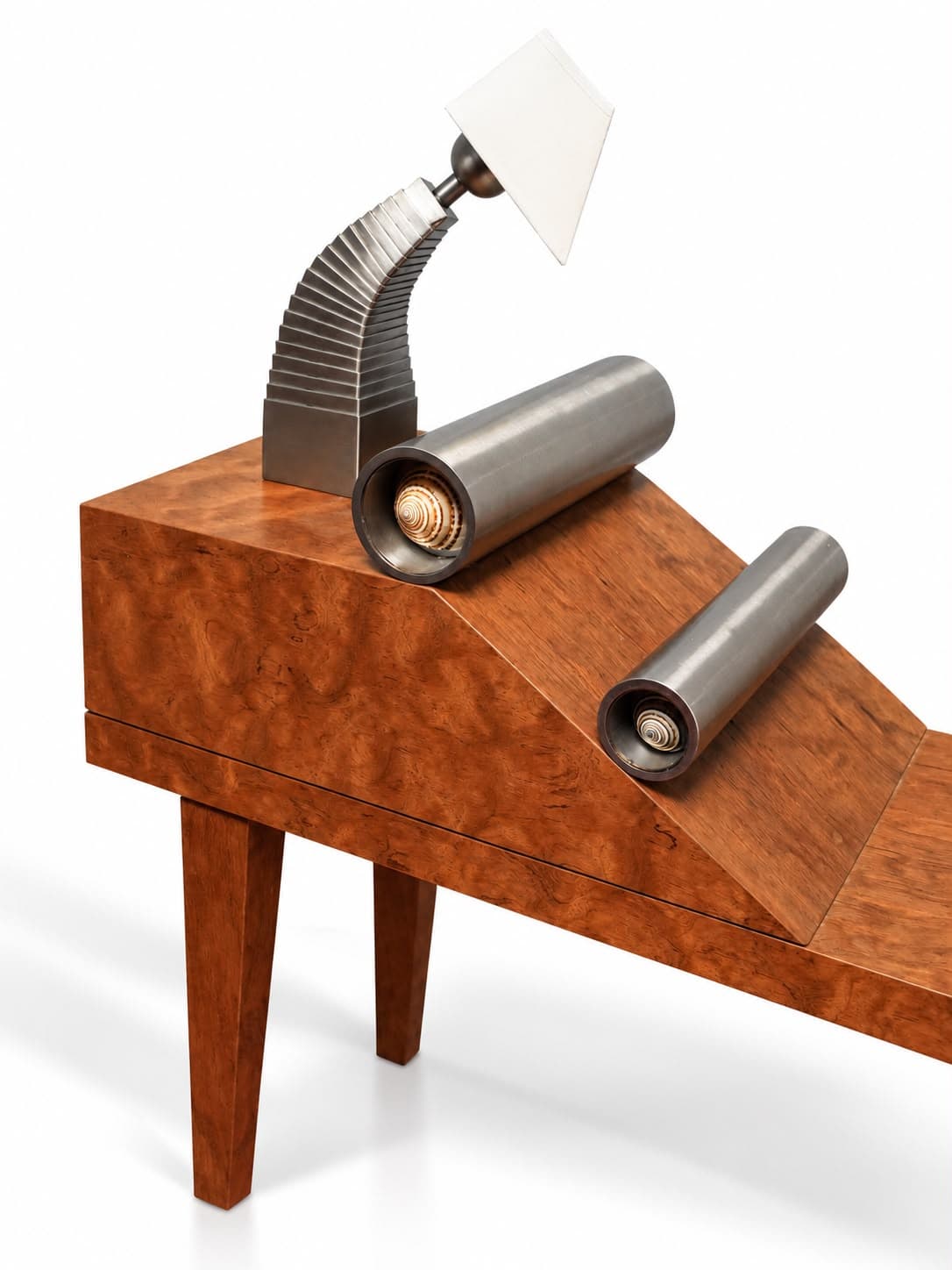 The head of the same bench close up: a fluted steel arm curving up to a white shade, and two steel cylinders resting on the burl ramp, each open at the near end to show a spiral seashell set inside it