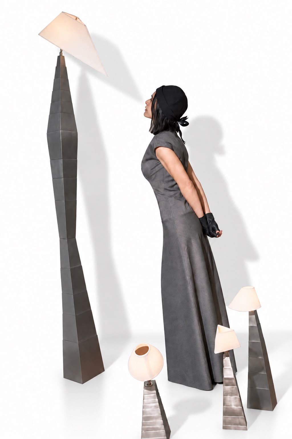 The artist standing in profile against a white cyclorama, chin raised and hands clasped behind her in a long grey dress and a black cap, among four of her stainless steel lamp totems — a tall leaning stack of blackened tapering blocks carrying a wide shade above her, and three shorter polished ones at her feet